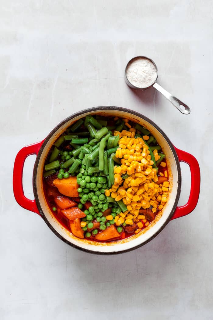 Vegetables including corn, green beans, and peas are shown on the surface of a pot of Mulligan stew.