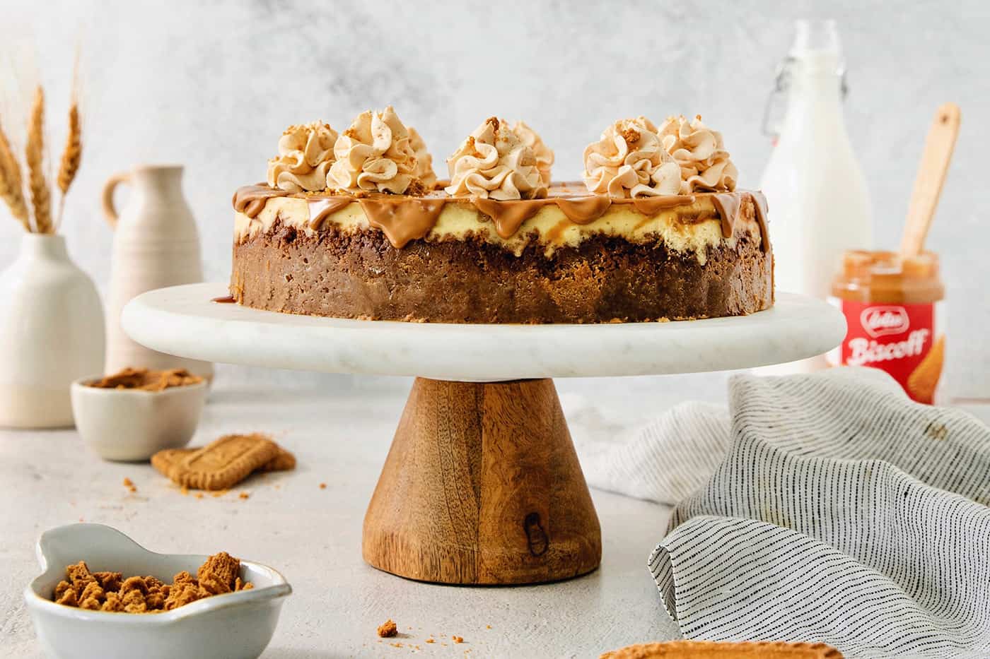 A biscoff cheesecake topped with butterscotch whipped cream is shown on a cake stand.