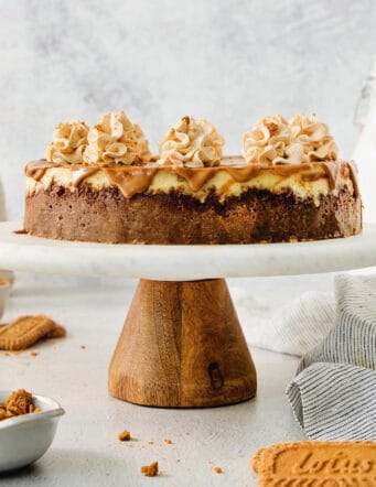 A biscoff cheesecake topped with butterscotch whipped cream is shown on a cake stand.