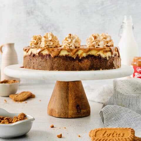 A biscoff cheesecake topped with butterscotch whipped cream is shown on a cake stand.