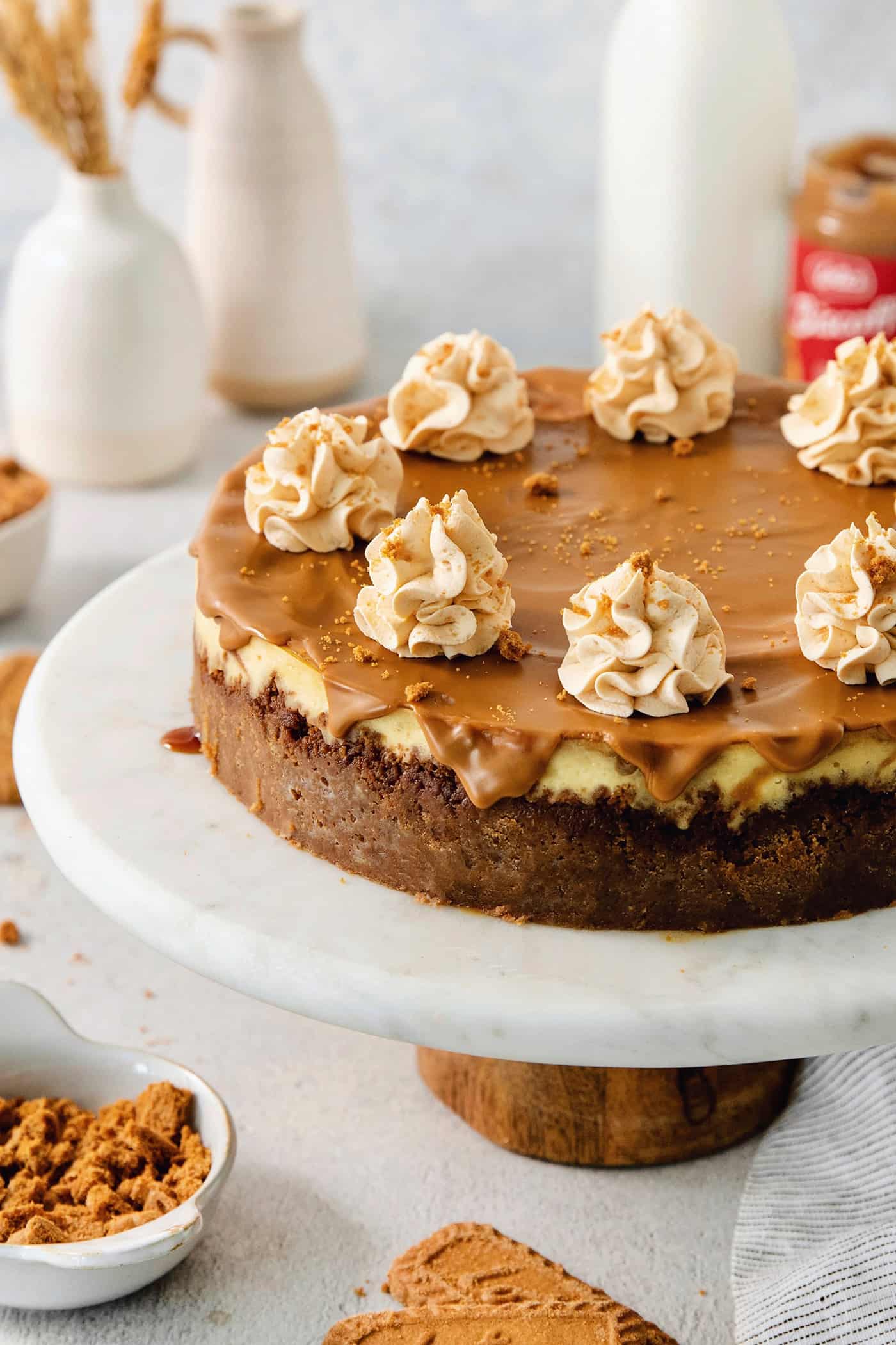 A biscoff cheesecake topped with butterscotch whipped cream is shown on a cake stand.