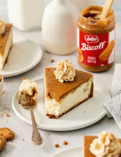 A slice of biscoff cheesecake is shown on a plate with biscoff cookie butter in the background.