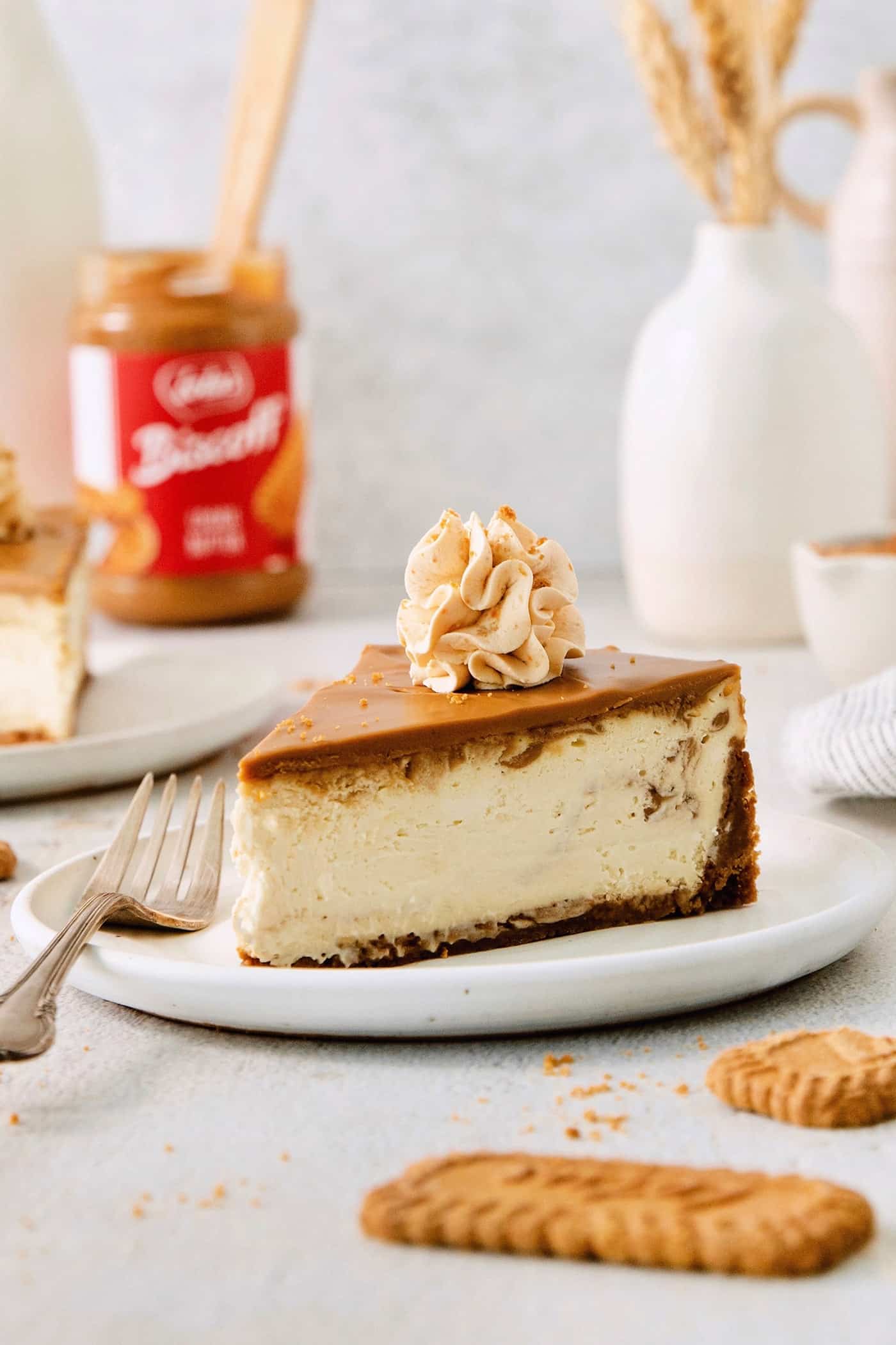 A slice of biscoff cheesecake is shown on a plate with biscoff cookie butter in the background.