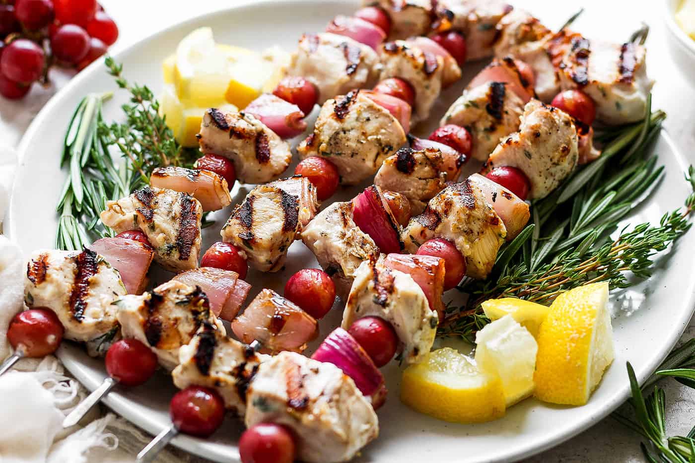 A white plate holds chicken kabobs with grapes on skewers with lemon next to them.