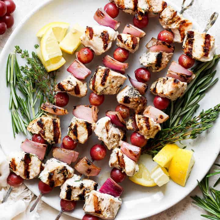 A white plate holds chicken kabobs with grapes on skewers with lemon next to them.