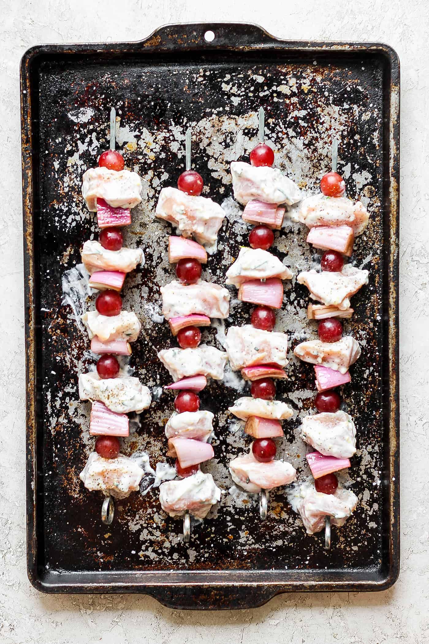 A baking sheet holds chicken kabobs with grapes before they are cooked.