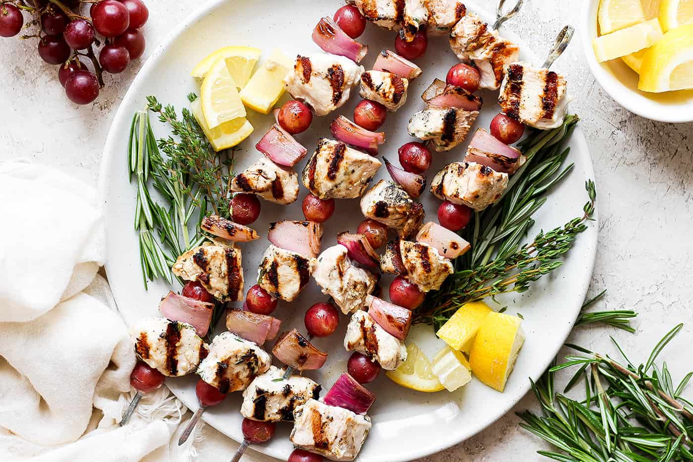 A white plate holds chicken kabobs with grapes on skewers with lemon next to them.