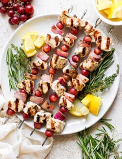 A white plate holds chicken kabobs with grapes on skewers with lemon next to them.