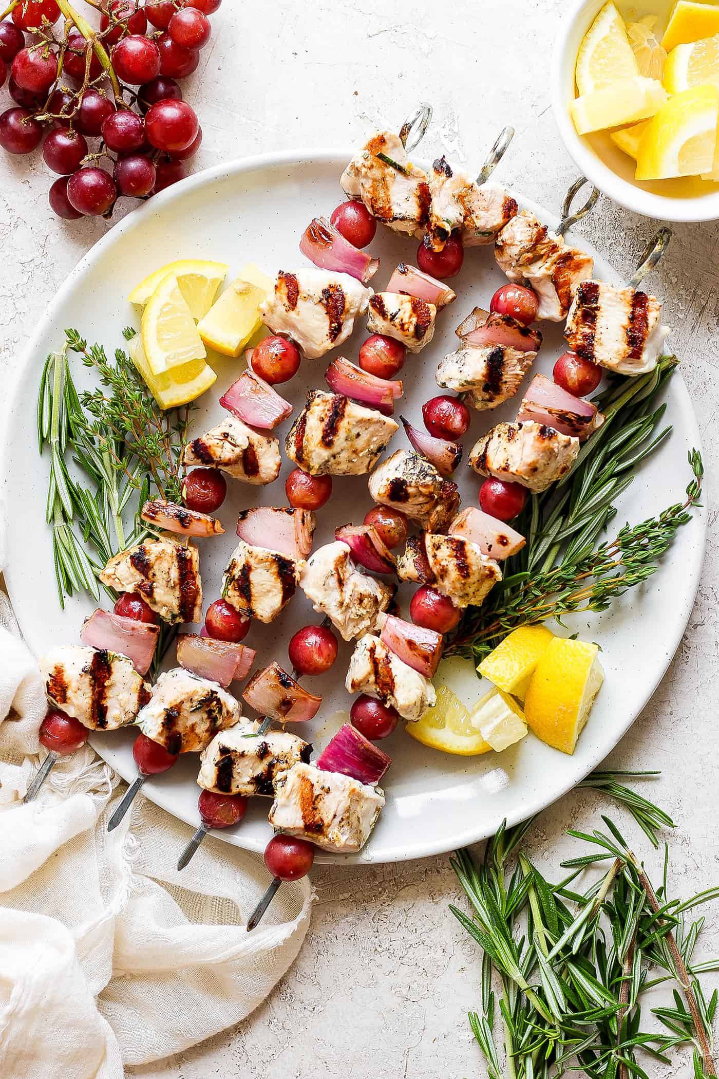 A white plate holds chicken kabobs with grapes on skewers with lemon next to them.