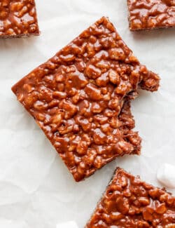 A chocolate rice krispie treat is shown with a bite taken out of it on a white background.