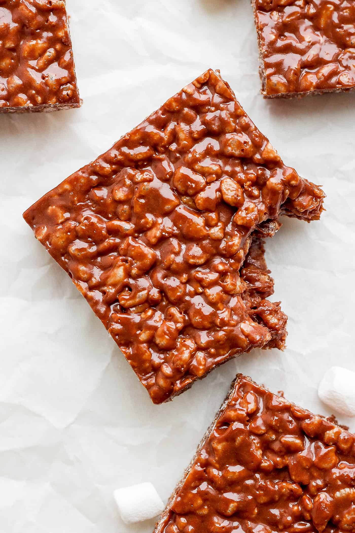 A chocolate rice krispie treat is shown with a bite taken out of it on a white background.