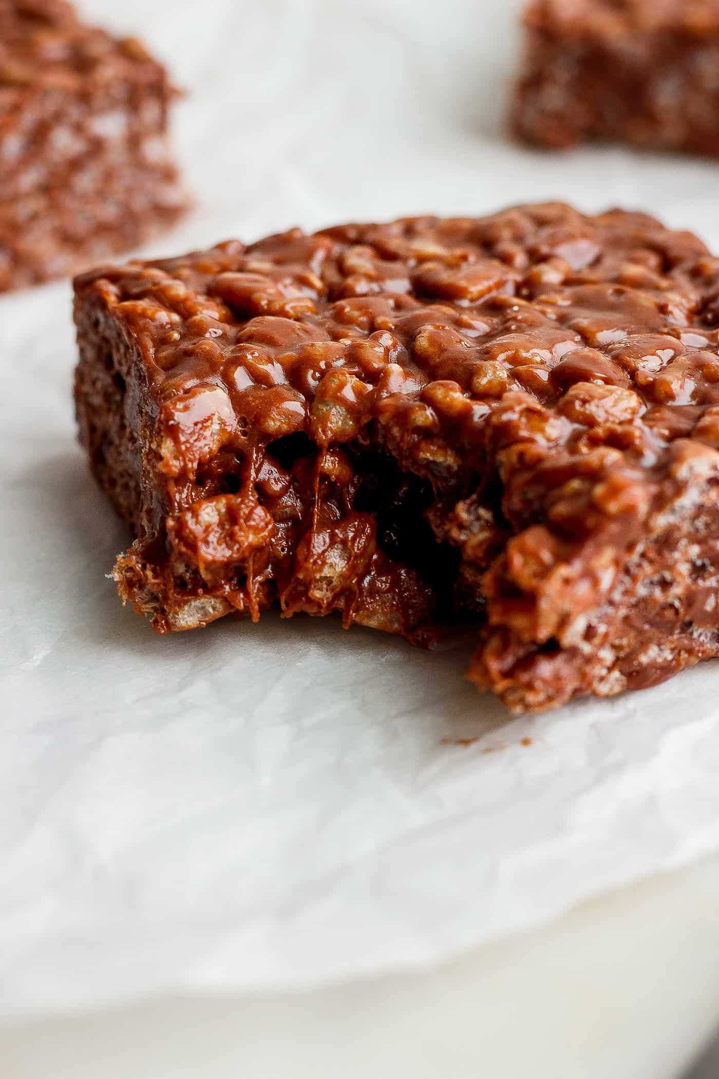 A chocolate rice krispie treat is shown with a bite taken out of it on a white background.