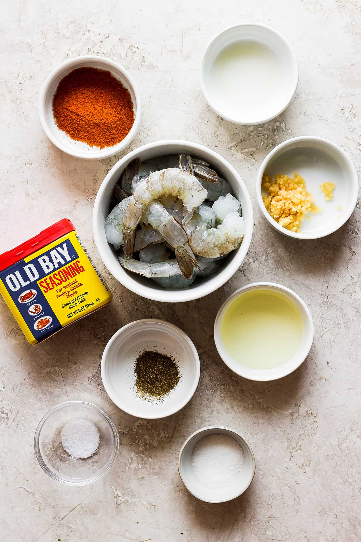 Ingredients needed for grilled shrimp are shown on a white background: shrimp, old bay seasoning, smoked paprika, olive oil, salt and pepper.