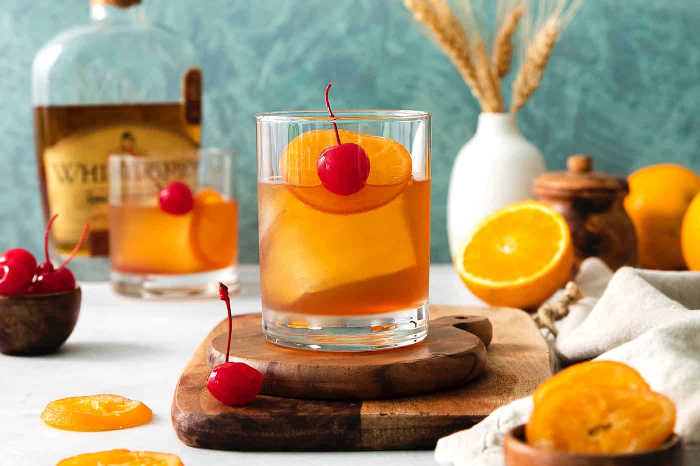 An old fashioned drink is shown on a wood board with more drinks and oranges in the background.