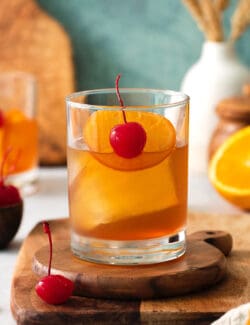 An old fashioned drink is shown on a wood board with more drinks and oranges in the background.