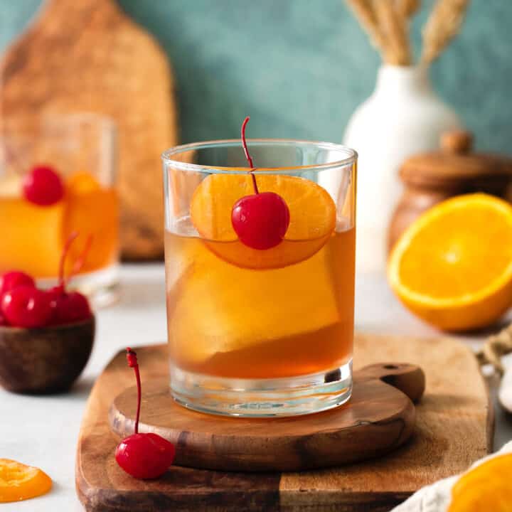 An old fashioned drink is shown on a wood board with more drinks and oranges in the background.
