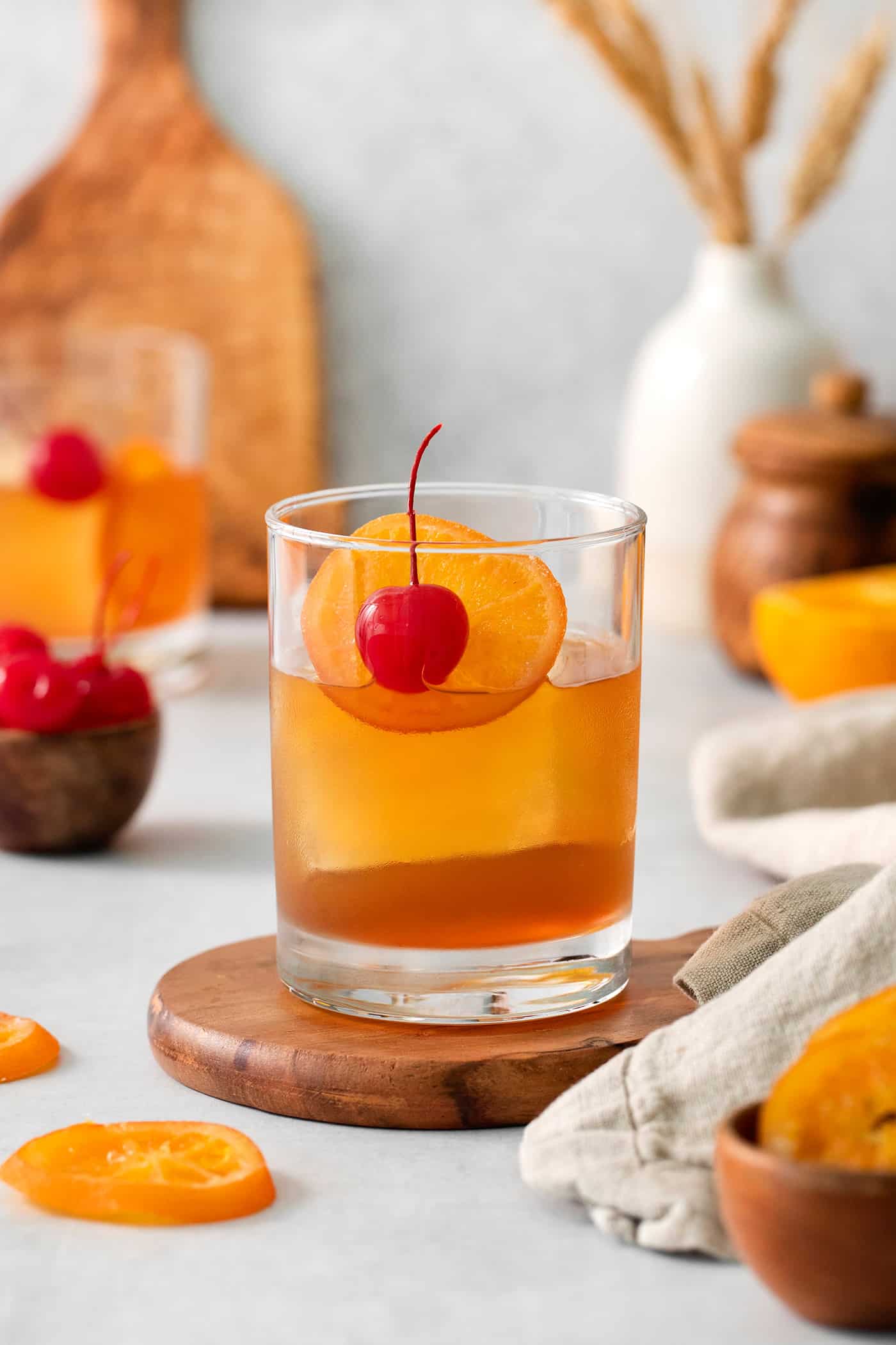 An old fashioned drink is shown on a wood board with more drinks and oranges in the background.