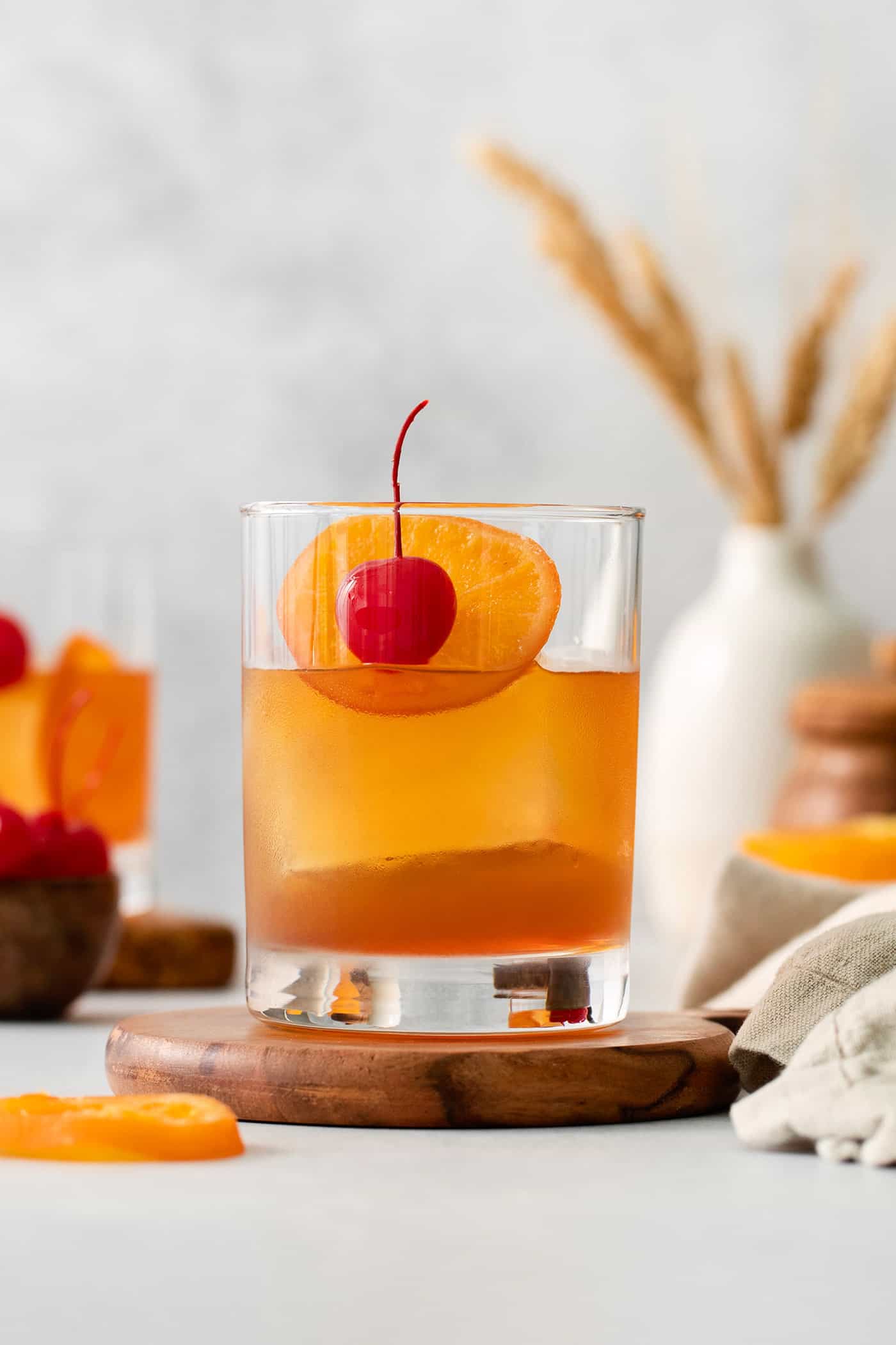 An old fashioned drink is shown on a wood board with more drinks and oranges in the background.