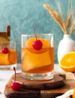 An old fashioned drink is shown on a wood board with more drinks and oranges in the background.