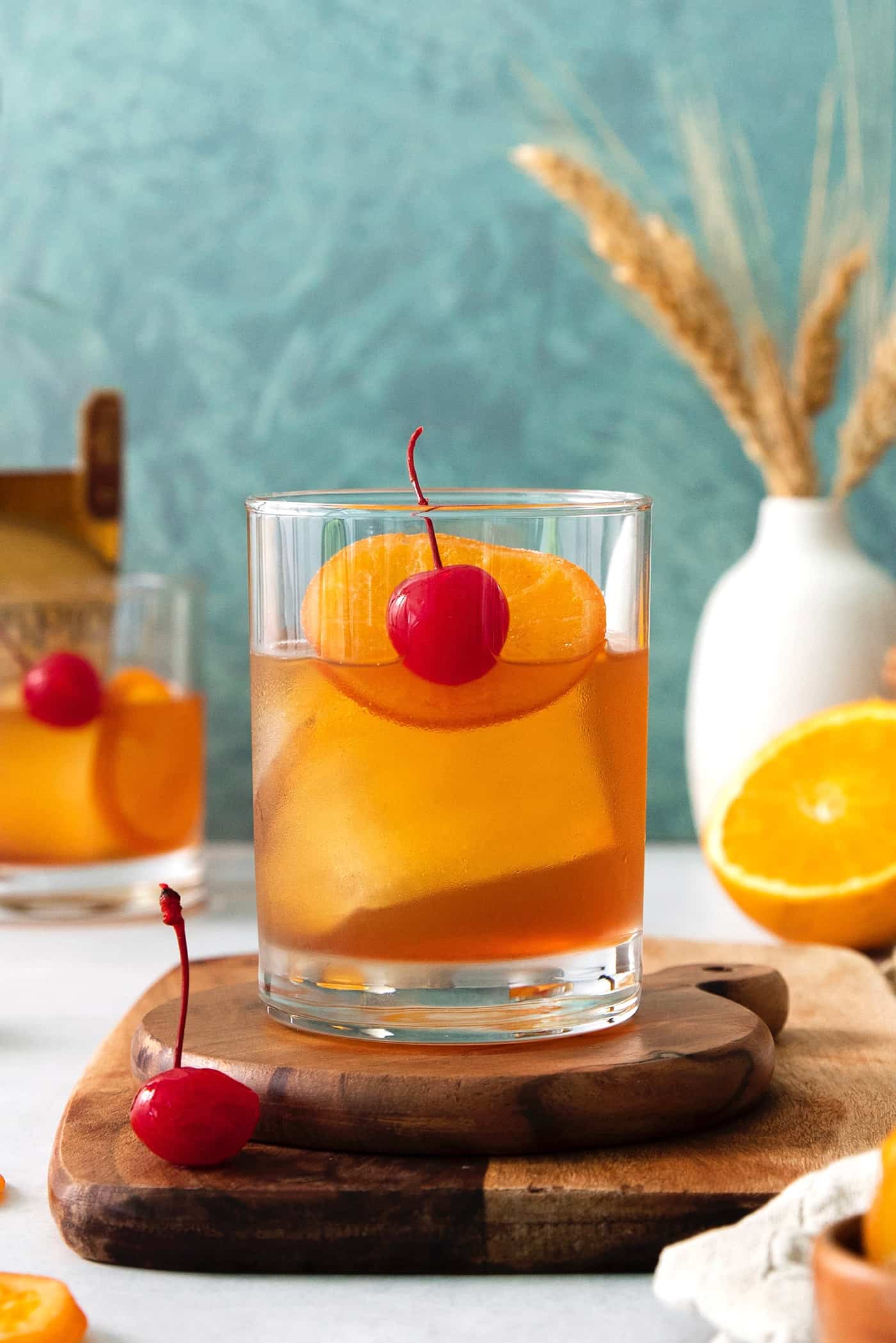 An old fashioned drink is shown on a wood board with more drinks and oranges in the background.