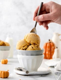 Scoops of orange pumpkin ice cream are shown in a white and gray bowl as a hand dips in a spoon.