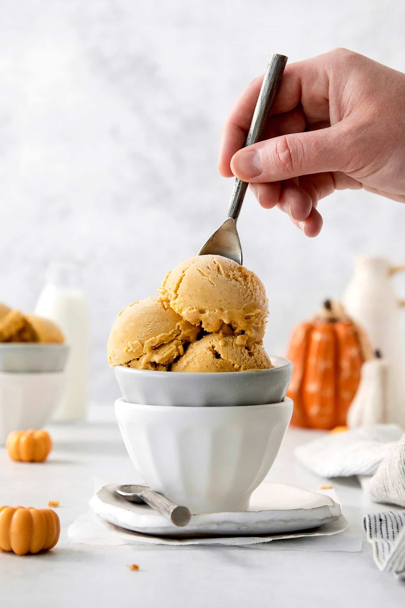 Scoops of orange pumpkin ice cream are shown in a white and gray bowl as a hand dips in a spoon.