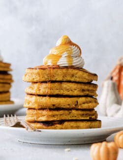 A stack of pumpkin pancakes topped with maple cinnamon whipped cream on a white plate.