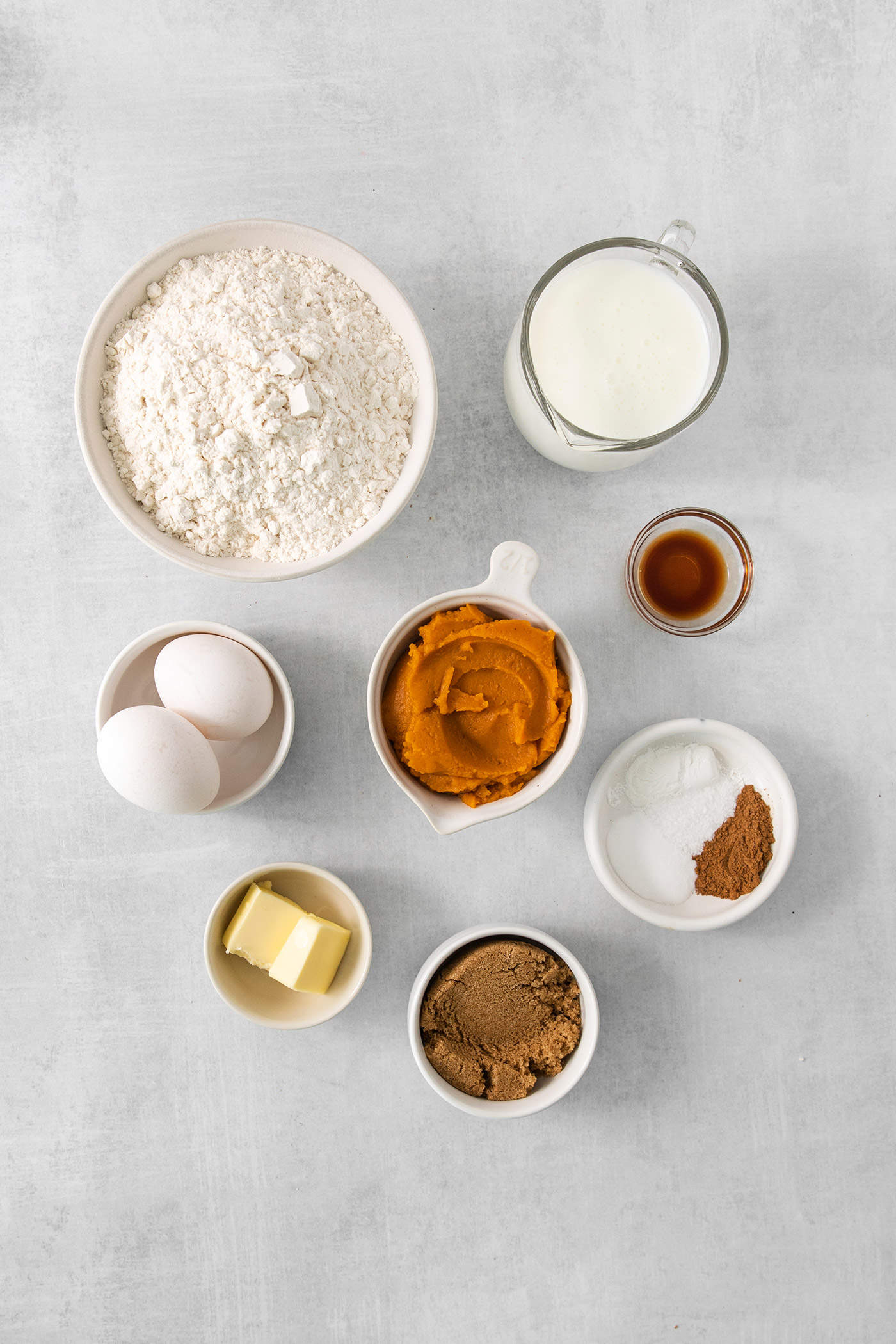 The ingredients for pumpkin pancakes are shown portioned into bowls, including flour, pumpkin puree, eggs, butter, pumpkin pie spice, vanilla, milk, and baking powder.