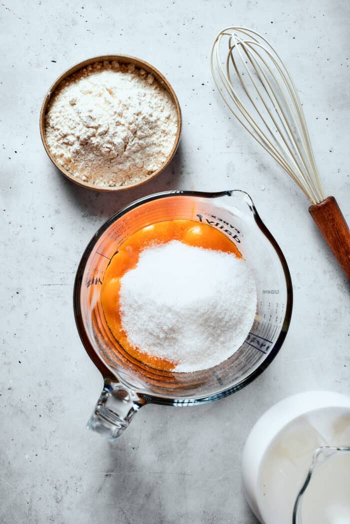 Eggs and sugar are shown in a glass measuring cup with a whisk next to it.