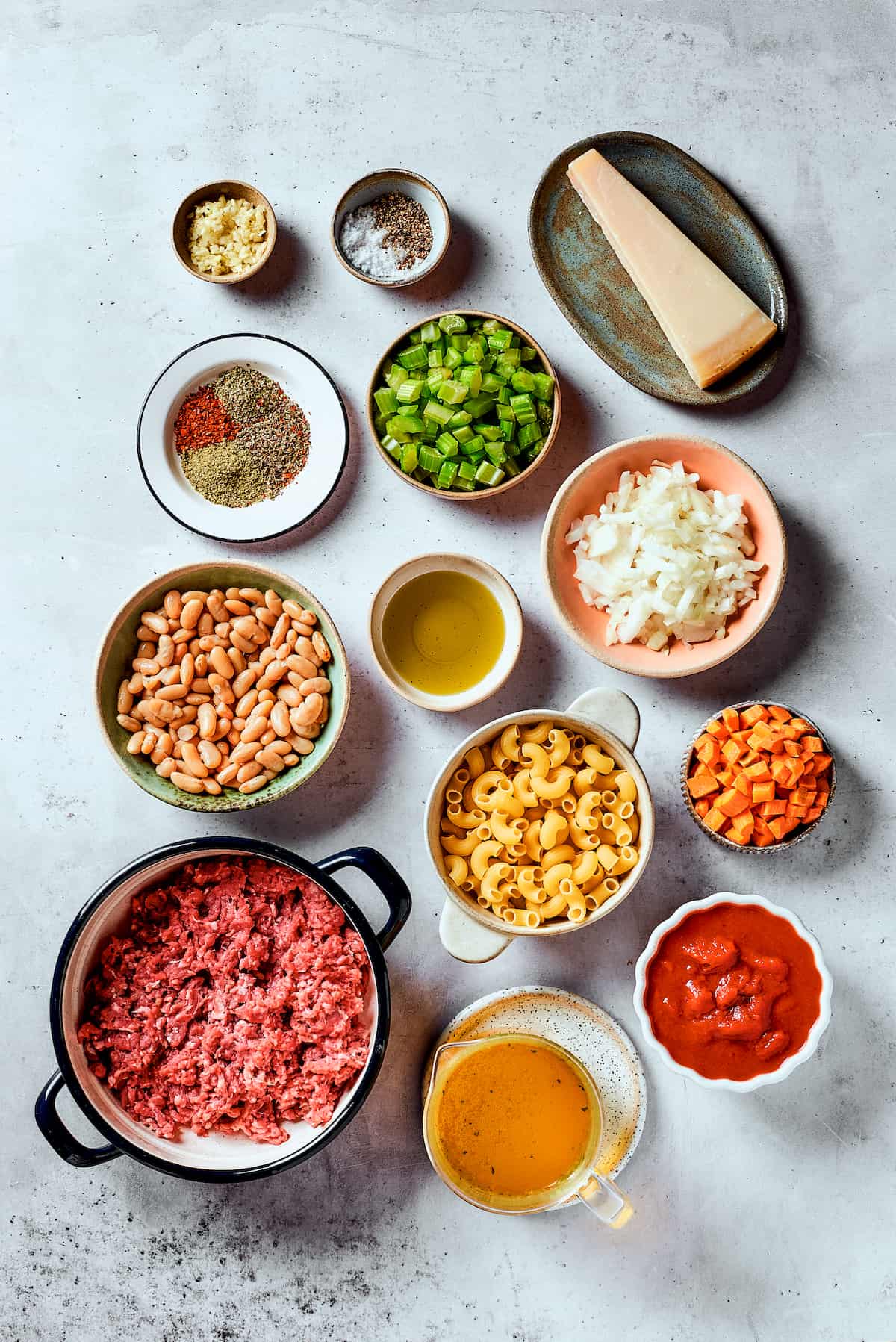Ingredients needed to make pasta fazool are shown portioned out on a white background: celery, carrots, pasta, meat, beans, tomatoes, garlic, onion, broth, herbs, and salt and pepper.
