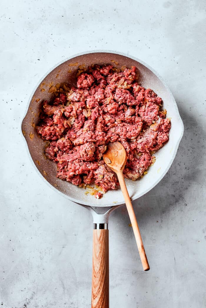 Meat cooks in a white skillet with a wooden spoon.