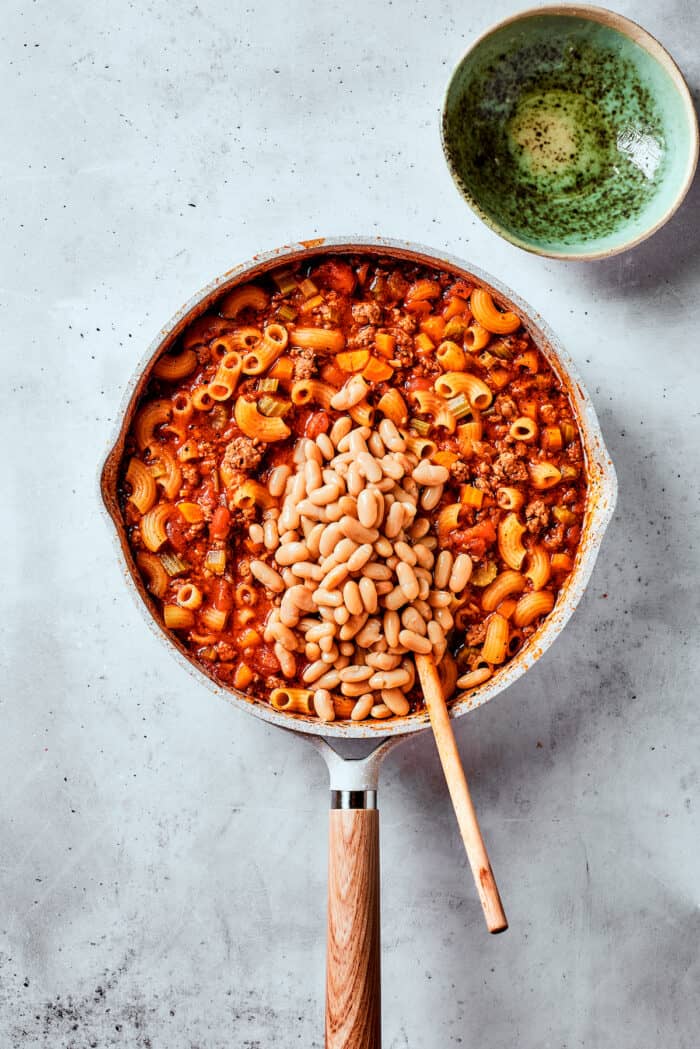 Beans are stirred into a pot of pasta fazool with a wooden spoon.