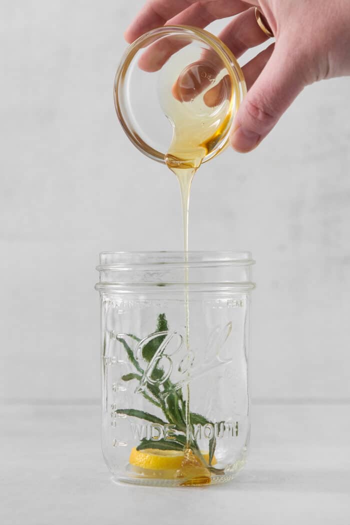 A hand pours honey into a mason jar with rosemary in it.