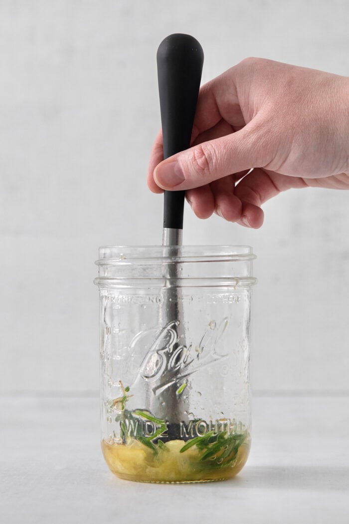 A hand muddles rosemary, lemon, and honey together in a glass jar.