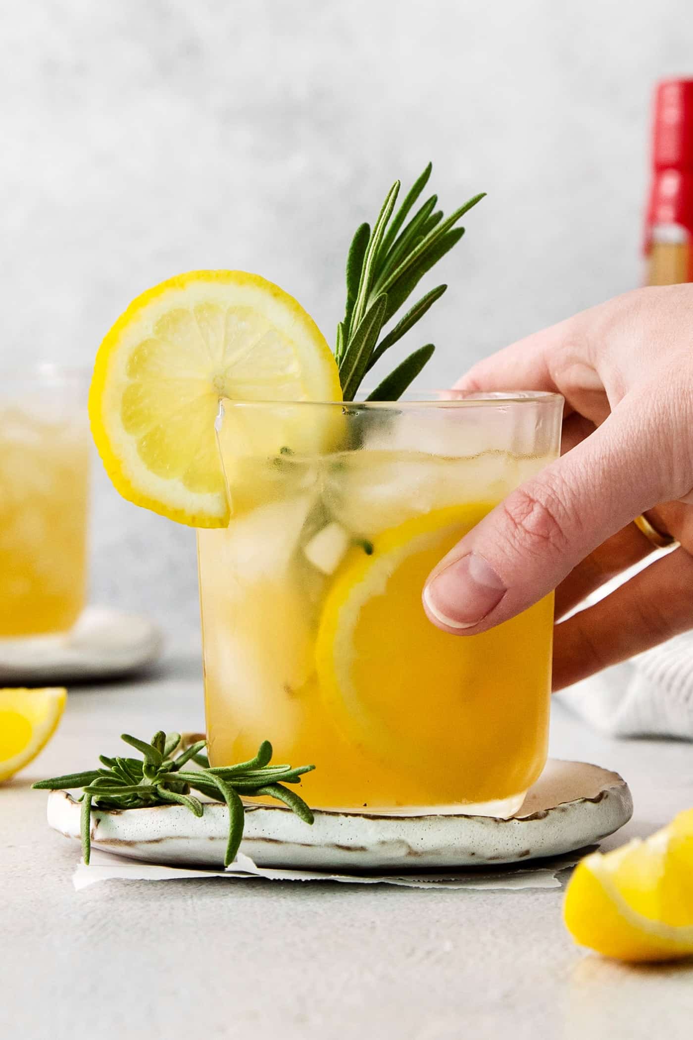 A hand holds a glass of bourbon limoncello cocktail toped with a lemon wheel and fresh rosemary.