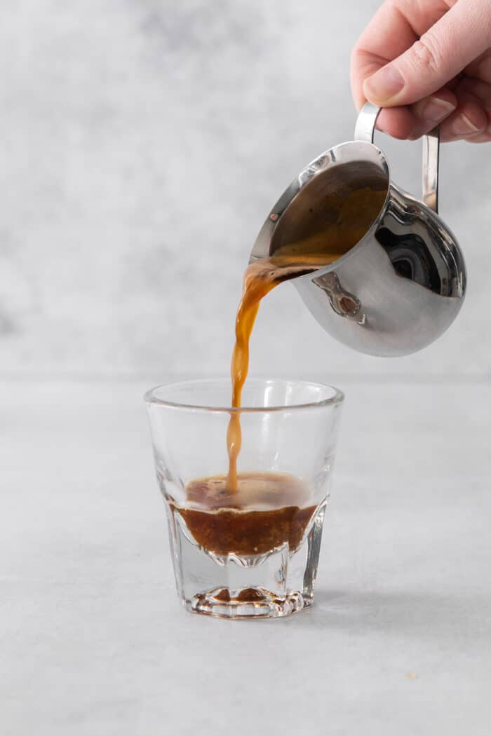 A hand pours a pitcher of espresso into a small glass.