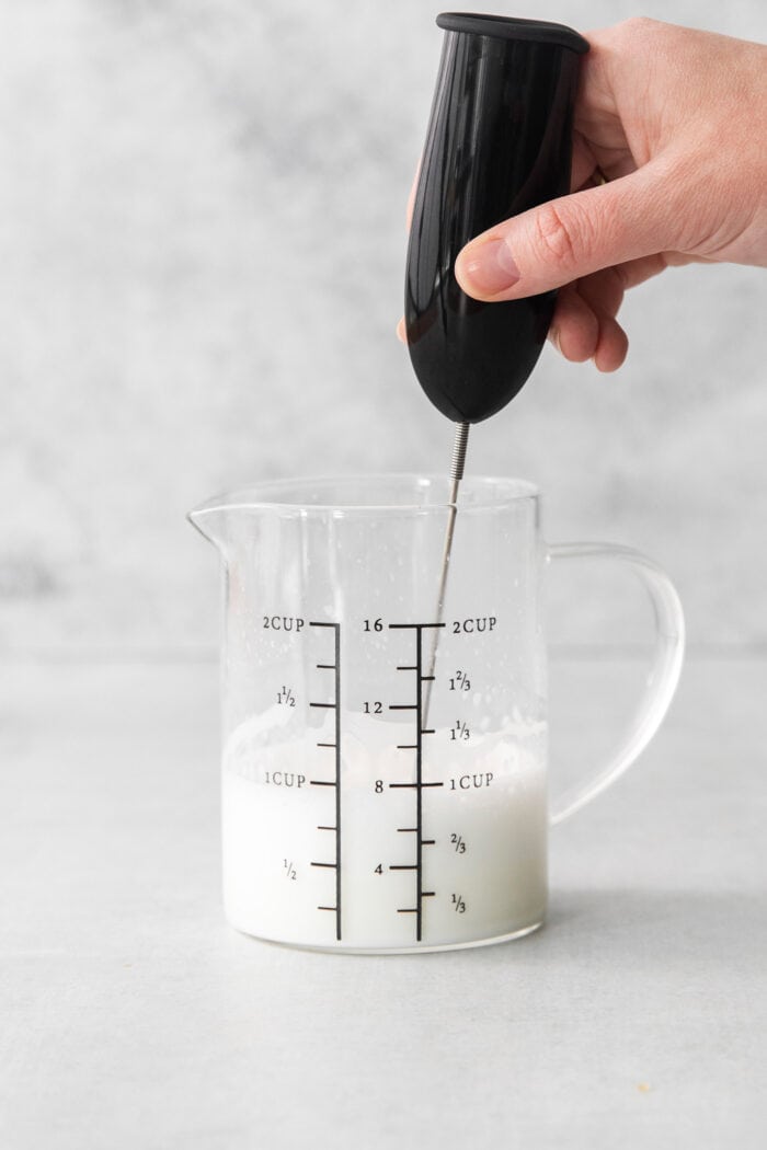 A hand holds a milk frother to froth a pitcher of milk.