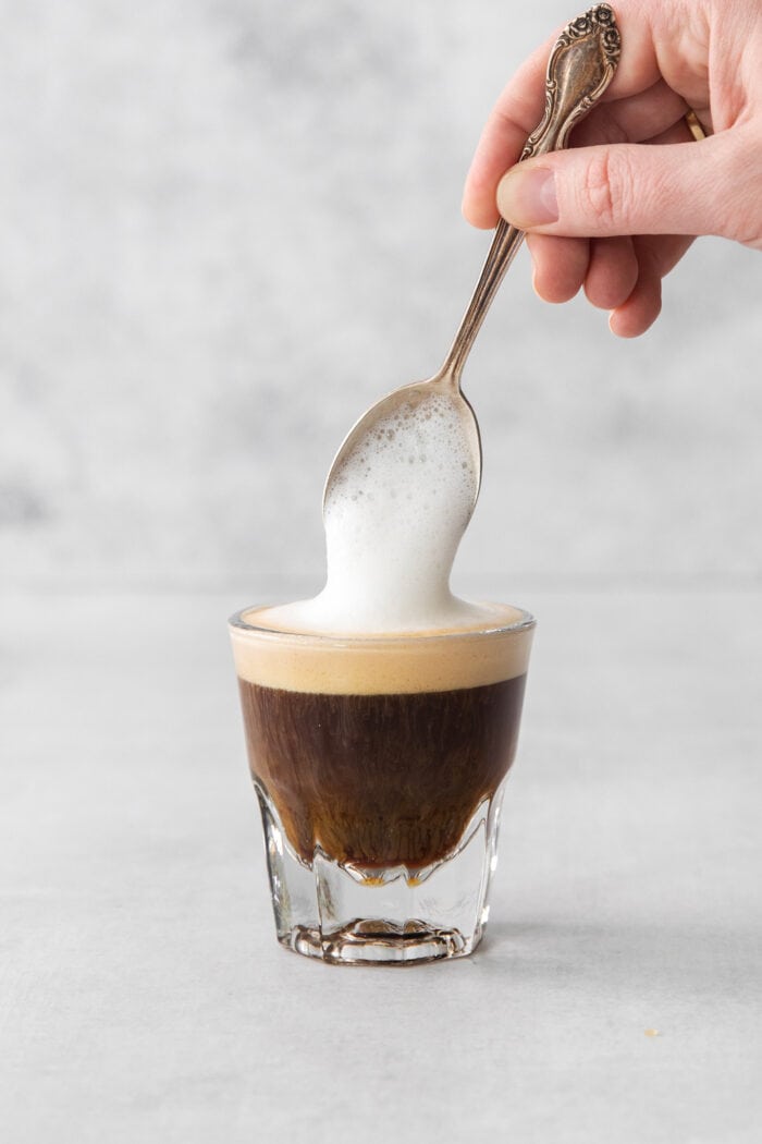 A hand holding a spoon tops a cortadito with frothed milk.