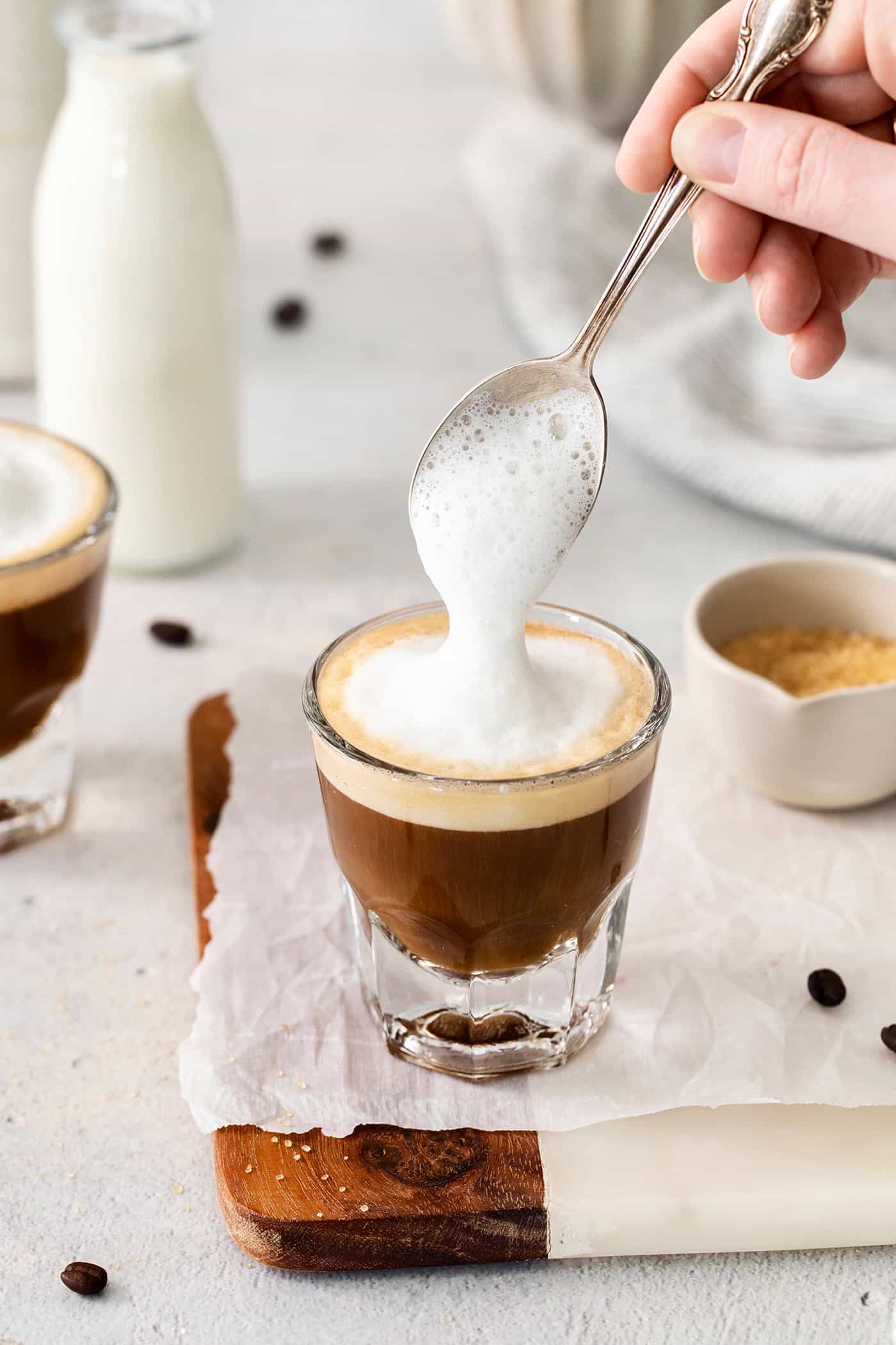 A hand holding a spoon tops a cortadito with frothed milk.