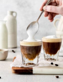 A hand holding a spoon tops cortaditos with frothed milk.