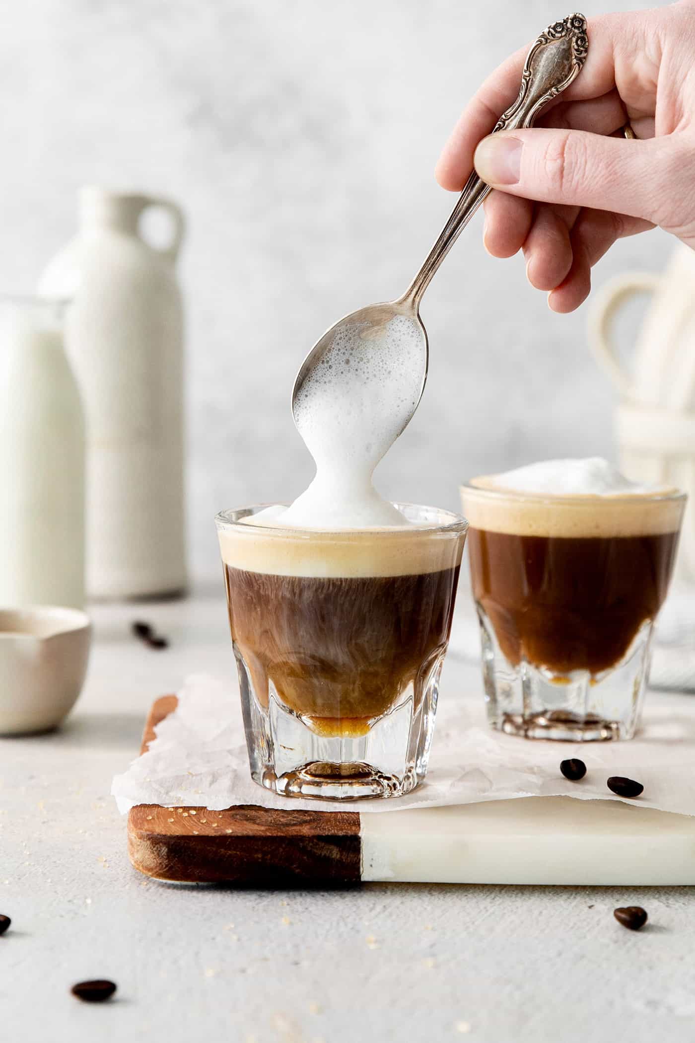 A hand holding a spoon tops cortaditos with frothed milk.