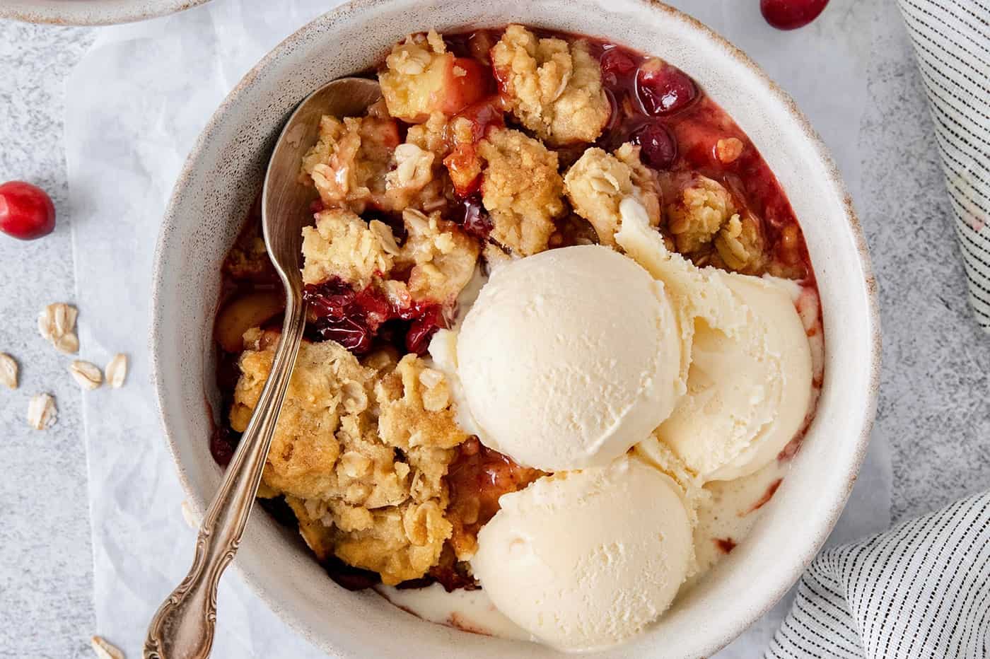 A white bowl of cranberry apple crisp with a spoon and topped with scoops of vanilla ice cream.