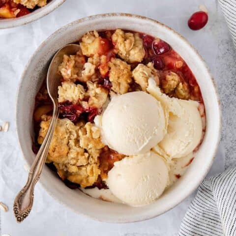 A white bowl of cranberry apple crisp with a spoon and topped with scoops of vanilla ice cream.