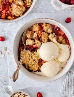 A white bowl of cranberry apple crisp with a spoon and topped with scoops of vanilla ice cream.