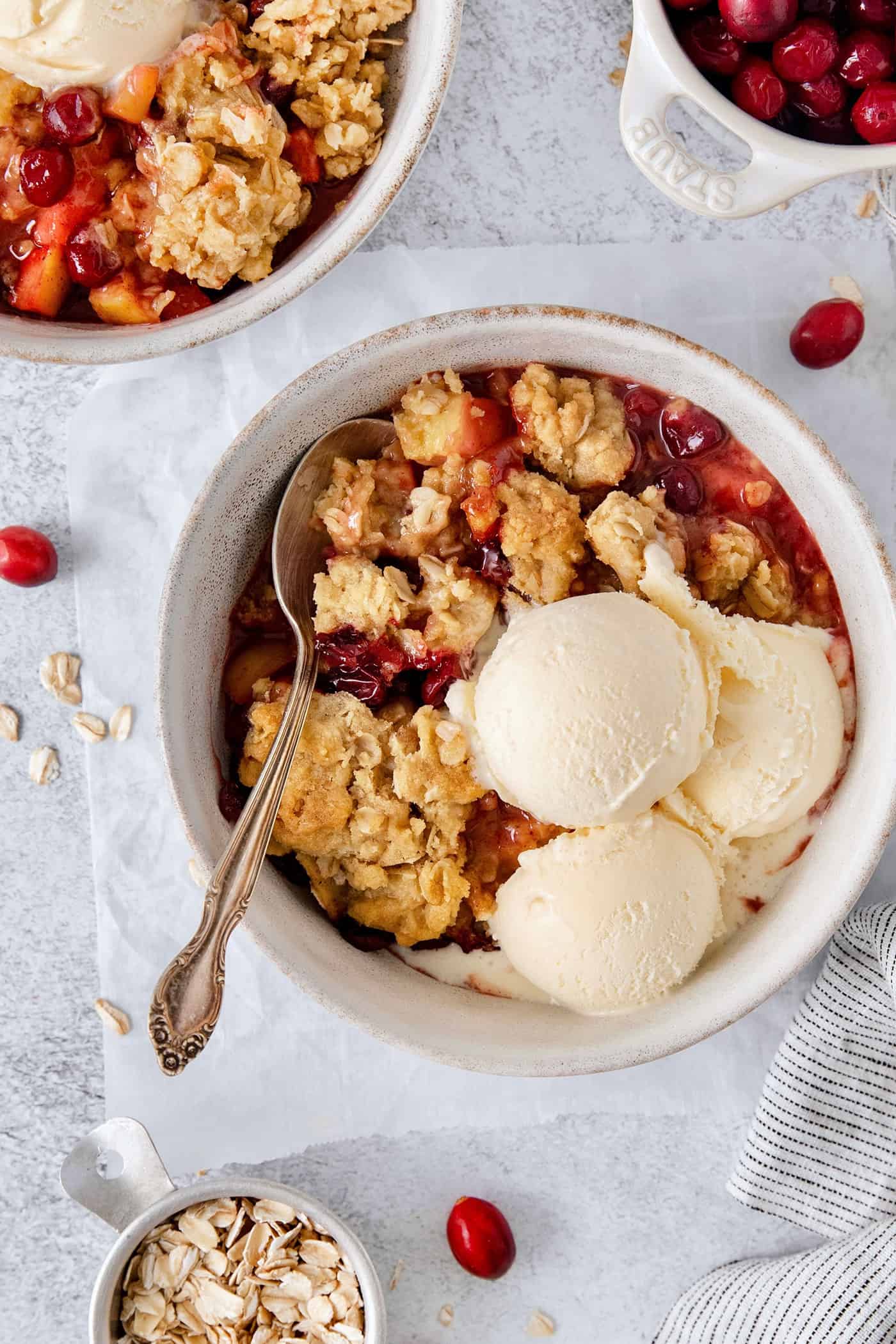 A white bowl of cranberry apple crisp with a spoon and topped with scoops of vanilla ice cream.