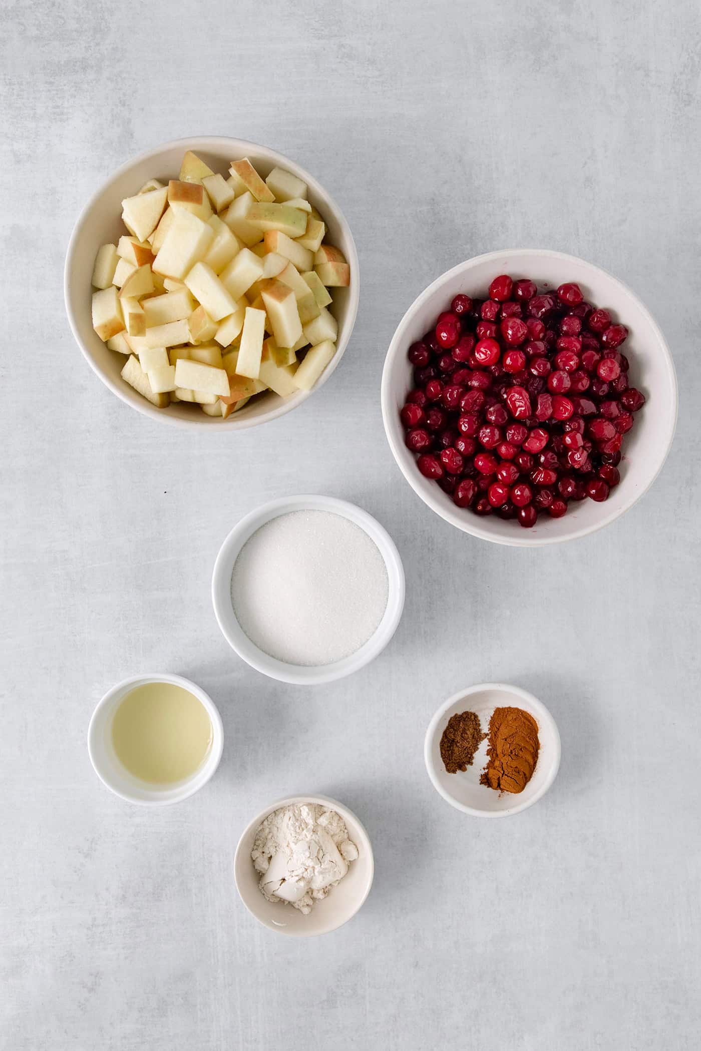 Ingredients for cranberry apple crisp filling are shown portioned out in bowls: cut apples, cranberries, sugar, spices, flour.
