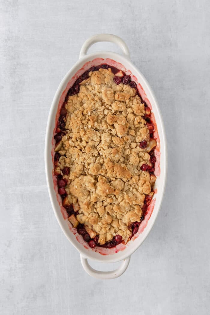 A white oval baking dish holds a baked cranberry apple crisp.