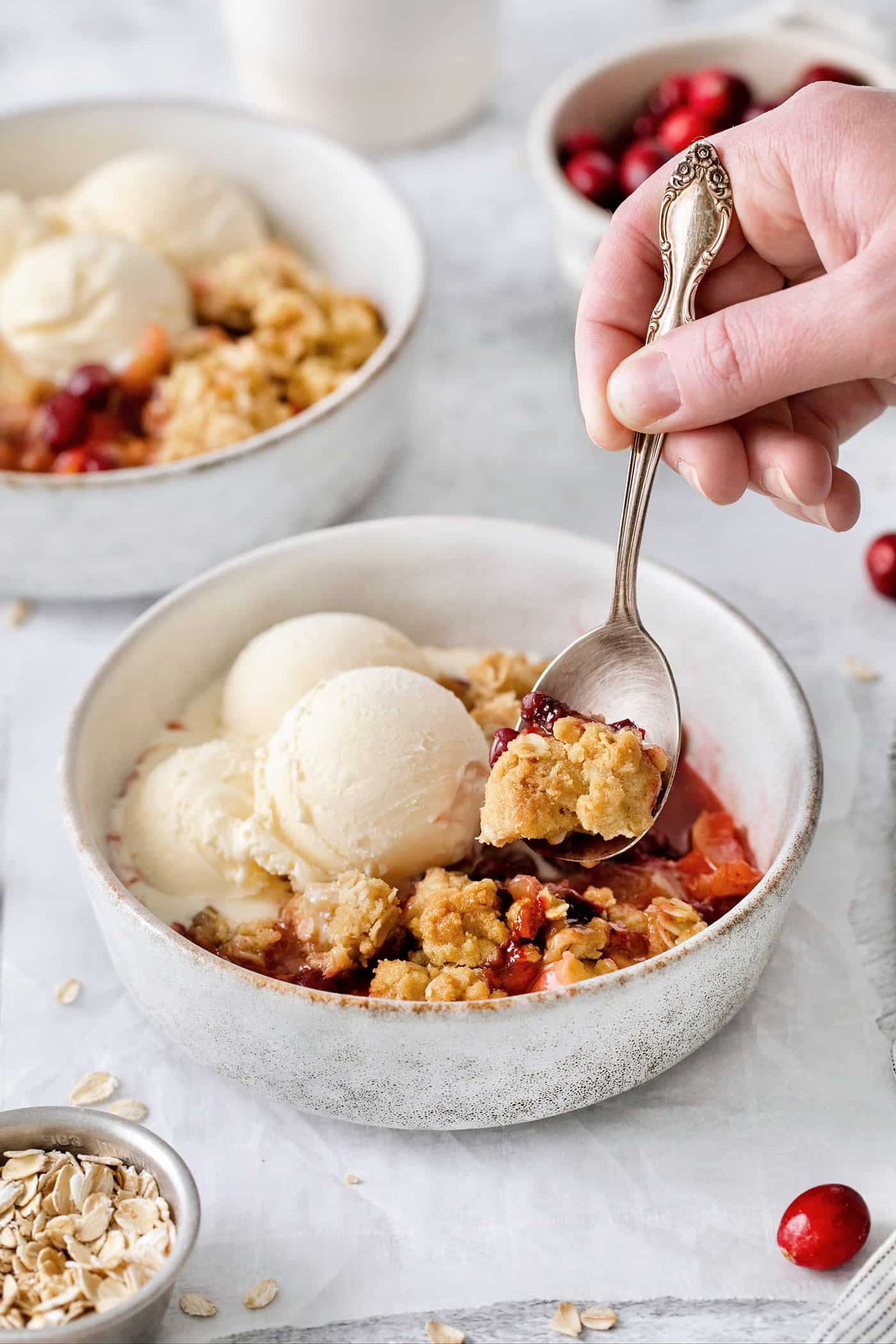 A hand holding a spoon dips into a bowl of cranberry apple crisp topped with vanilla ice cream.