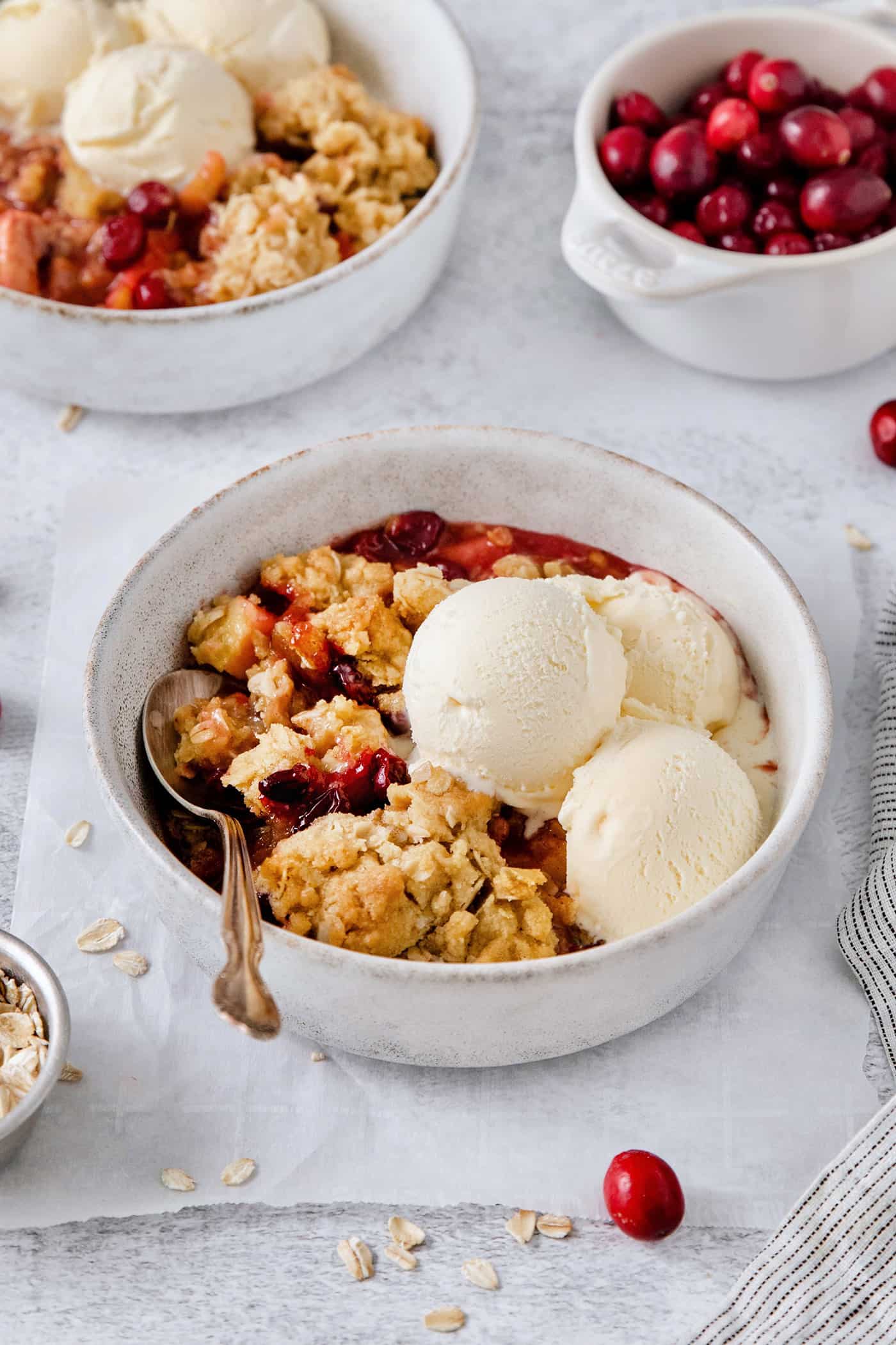 A white bowl of cranberry apple crisp with a spoon and topped with scoops of vanilla ice cream.