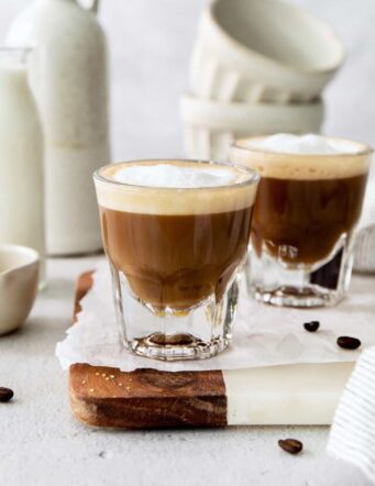 Two cortaditos are shown on a board with milk in the background.